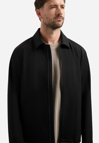 Mann mit kurzer Behaarung, der eine schwarze Jacke mit Reißverschluss über einem beigen Hemd trägt und leicht nach rechts blickt, vor einem schlichten weißen Hintergrund.