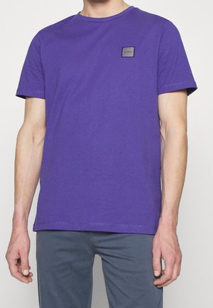 Mies, jolla on yksivärinen violetti lyhythihainen T-paita, jossa on pieni BOSS-logo vasemmalla rinnassa, ja harmaat housut.