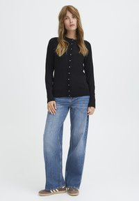 Cárdigan negro de manga larga con botones, combinado con jeans de pierna ancha azul claro. El atuendo incluye zapatos marrones con rayas blancas.