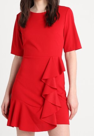 Femme portant une robe rouge à manches courtes avec des volants asymétriques sur le devant et l'ourlet.