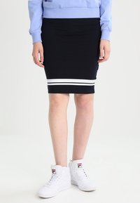 Osoba v černé sukni se dvěma bílými pruhy u spodního okraje, světlomodrém svetru a bílých teniskách s vysokým střihem a logem.