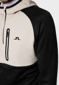 Sweat à capuche avec un blocage de couleurs crème et noir, comportant une poche zippée à l'avant et un petit logo. Texture lisse et coupe sportive.