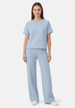 Nainen seisoo ja hänellä on vaaleansininen lyhythihainen paita ja siihen sopivat leveälahkeiset housut sekä valkoiset lenkkarit yksiväristä taustaa vasten.