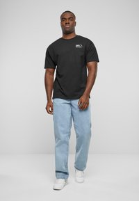 Sort bomulds-T-shirt med korte ærmer, prydet med et lille logo i grøn og blå på venstre side. Sammen med lyseblå jeans og hvide sneakers.