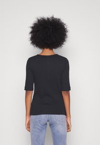 Person med lockigt hår som bär en svart t-shirt med halv ärm och ljusblå jeans, stående vänd mot en enkel vit vägg.