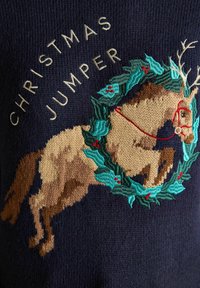 Blauer gestrickter Weihnachten-Pullover mit einem bestickten Pferd, das mit einem Kranz aus Stechpalme und Beeren geschmückt ist, akzentuiert mit goldener Schrift "WEIHNACHTS-PULLOVER".