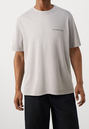 Mand iført lysegrå kortærmet t-shirt med lille "Abercrombie and Fitch"-logo på venstre bryst og mørke bukser.