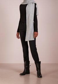 Schwarzes geripptes Langarmoberteil, maßgeschneiderte schwarze Hose, grauer gestrickter Schal drapiert und schwarze Leder-Cowboystiefel vor einem neutralen Hintergrund.