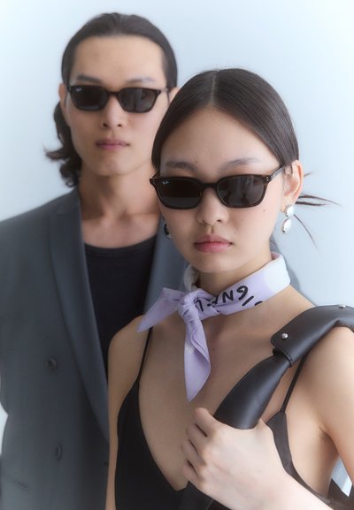 Lunettes de soleil avec des verres foncés et une monture rectangulaire. L'arrière-plan présente une couleur claire et lisse. Le modèle porte une robe noire et une écharpe violet clair.