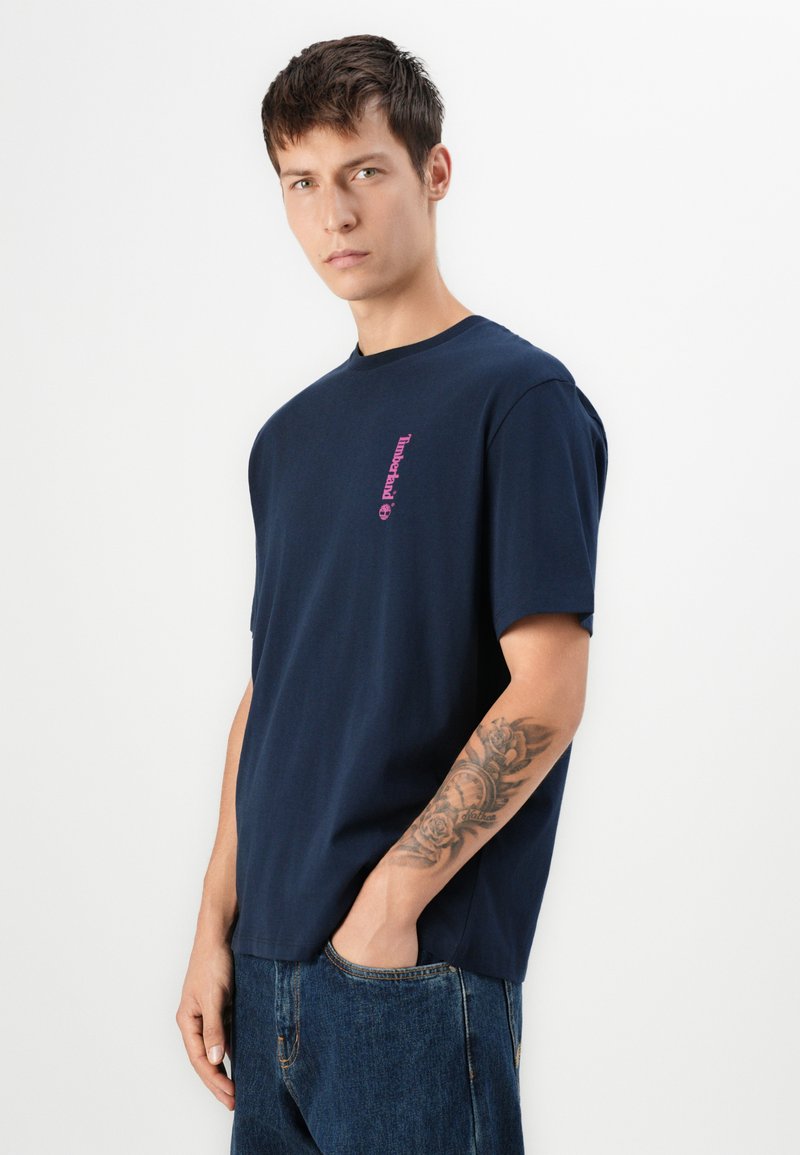 Młody mężczyzna w granatowej koszulce Timberland i dżinsach, pokazujący szczegółowy tatuaż z kwiatami i zegarem na lewym przedramieniu.