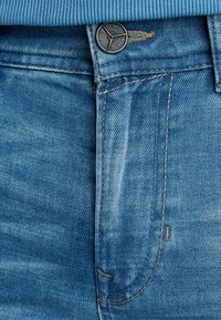 Pohled zblízka na modré džíny s knoflíkem označeným jako "Legend Jeans" a podrobným šitím kolem pasu a zipu.