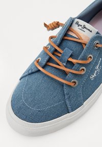 Sapatilhas de denim azul com atacadores de couro, sola de borracha branca e ilhós circulares. Apresenta o logotipo na lateral e uma superfície de tecido texturizado.