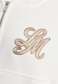 Material alb cu litere "F" și "M" bej brodate și împletite lângă un fermoar argintiu pe un articol vestimentar, probabil un hanorac sau o jachetă.