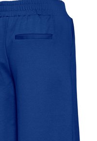 Zadnji pogled na modre hlače z elastičnim pasom in eno razporko na desni strani.
