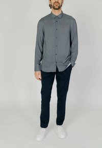 Camicia button-up in un tessuto grigio-blu con un motivo geometrico; taglio slim; abbinata a pantaloni scuri e sneakers bianche.