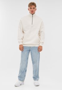 Light beige zip-up sweatshirt with a high collar, featuring a front pocket and ribbed cuffs, paired with loose-fitting light blue jeans and white sneakers.