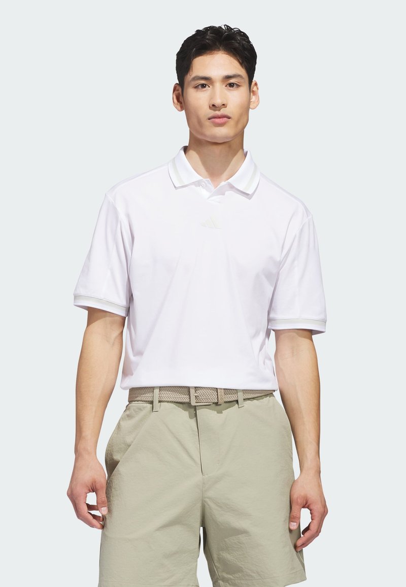 Λευκό πόλο πουκάμισο με κοντά μανίκια, επίπεδη γιακά και διακριτικό λογότυπο στο στήθος, συνδυασμένο με ανοιχτό καμήλι σορτς και πλεκτή ζώνη.