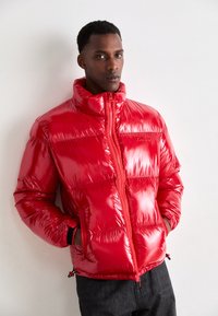 Red puffer jacket with a glossy finish, high collar, zip closure, and front pockets. Features elastic cuffs and adjustable hem for fit.