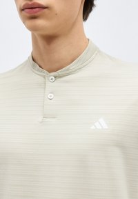 Άτομο που φορά ένα ανοιχτό μπεζ ριμπ πλεκτό πόλο με μανταρίνι γιακά, δύο κουμπιά και λευκό λογότυπο Adidas στο στήθος.