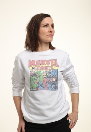 Kvinde med skulderlangt brunt hår iført en hvid sweatshirt med Marvel Comics-figurer, stående med den ene hånd på hoften.