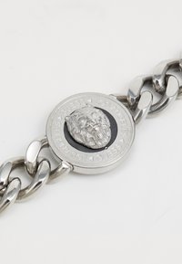 Bracelet en métal argenté avec une chaîne, mettant en avant un pendentif circulaire avec une tête de lion en relief et l'inscription gravée "GUESS LOS ANGELES".