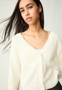 Weißer Strickcardigan mit V-Ausschnitt, der fünf cremefarbene Knöpfe und gerippte Textur aufweist. Glatte Innenschicht, die über einem weißen Oberteil getragen wird.