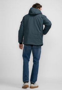 Mørkeblå puffjakke med hætte, glat tekstur, elastiske manchetter og en afslappet pasform. Bæres med blå jeans og hvide sneakers.