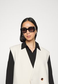Óculos de sol oversized pretos com lentes escuras, combinados com um colete branco sem mangas sobre uma camisa preta. Acessório prateado nos óculos.