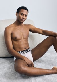 Λευκά εσώρουχα Calvin Klein με μαύρη ελαστική ζώνη, με έντονες λευκές επιγραφές. Μοντέλο καθισμένο σε ένα μαλακό, υφασμένο χαλί.