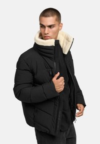 Melna pūkaina jaka ar augstu apkakli, kurai ir krēma krāsas vilnas oderējums, priekšējais rāvējslēdzējs un sānu kabatas. Gluda tekstūra ar quilted rakstu.