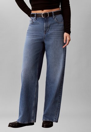 Persoon die hooggetailleerde wijde blauwe spijkerbroek draagt met zwarte riem, zwart cropped top met lange mouwen en zwarte schoenen, staand tegen een grijze achtergrond.