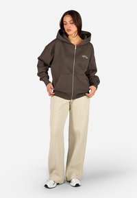 Brauner Kapuzenpullover mit Reißverschluss aus weichem Material, mit einem Frontlogo und seitlichen Taschen, kombiniert mit cremefarbigen weiten Hosen.