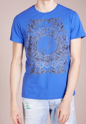 T-shirt en coton bleu avec un design graphique noir orné de motifs élaborés et un emblème central portant l'inscription "VERSACE JEANS". Manches courtes, col rond.