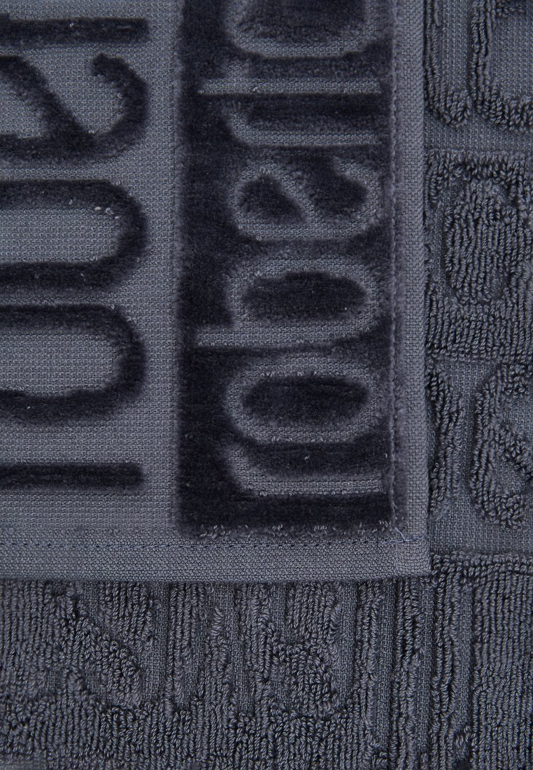 Dunkelgraues Baumwollhandtuch mit einer strukturierten Oberfläche, die erhabene Schriftzüge und Muster aufweist. Die Ränder sind mit einer glatten Einfassung versehen.