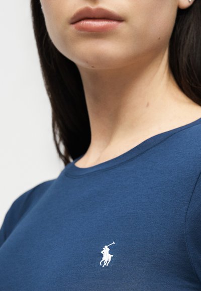 Γυναίκα που φοράει μπλούζα μπλε, λαιμόκοψης crew-neck, με λευκό κεντημένο λογότυπο παίκτη πόλο στο στήθος, εμφανίζοντας το κάτω μέρος του προσώπου και τον λαιμό.