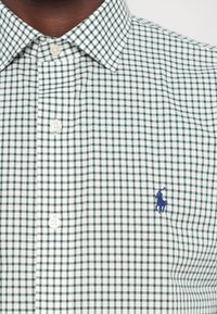 Zeleno-biely károvaný košeľový vrch s golierom na gombíky, s malým modrým vyšitým logom na ľavej hrudi, vyrobený z tkaninového materiálu.