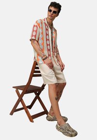 Kolorowa, wzorzysta, krótko rękawowa koszula w pomarańczowe, czerwone i kremowe diamenty, zestawiona z beżowymi szortami i kamuflażowymi klapkami na drewnianym krześle.