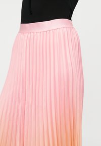 Frau trägt einen hoch taillierten, plissierten Rock mit rosa bis orangefarbenem Ombre-Verlauf und ein schwarzes Oberteil vor einem hellen Hintergrund.