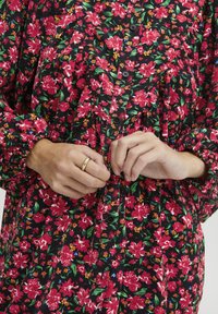 Blusa floreale nera con motivi rosa e verdi. Presenta polsini tondi, chiusura con bottoni e tessuto morbido e leggero. Anello dorato sulla mano.