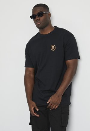 Black cotton t-shirt featuring a round logo with gold accents on the left chest. Model wears black sunglasses, paired with dark cargo pants.