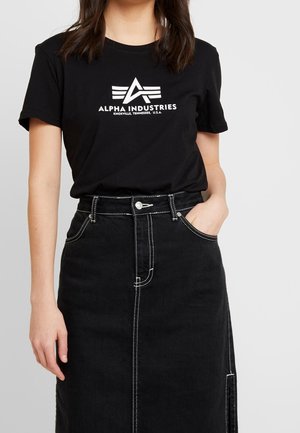 Kvinde med mørkt hår iført en sort Alpha Industries t-shirt og højtaljet sort nederdel med hvide syninger, den ene hånd i lommen.