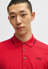 Rød polo shirt lavet af tekstureret stof med en sort krave og sorte detaljer. Tre-knap lukning med logo broderet på brystet.