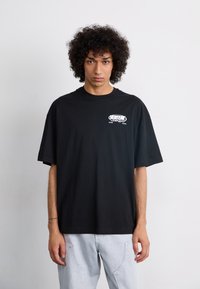 Sort t-skjorte i bomull med korte ermer, med et lite hvitt grafisk motiv på brystet. Kombinert med lyseblå denim-bukser.