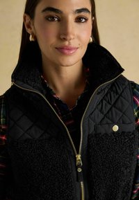 Czarna pikowana kamizelka z detalami z sztucznego futra, złotym zamkiem błyskawicznym i zatrzaskami, nałożona na wielokolorową koszulę w kratę.