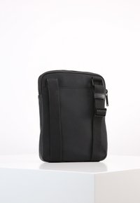 Borsa a tracolla rettangolare nera con tracolla regolabile e zip superiore, posizionata in verticale su una superficie bianca contro uno sfondo semplice.