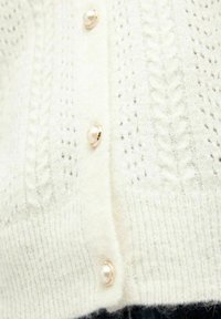 Cardigan en tricot blanc avec un motif texturé, présentant des boutons semblables à des perles et des détails subtils. Tissu doux et vue en gros plan.