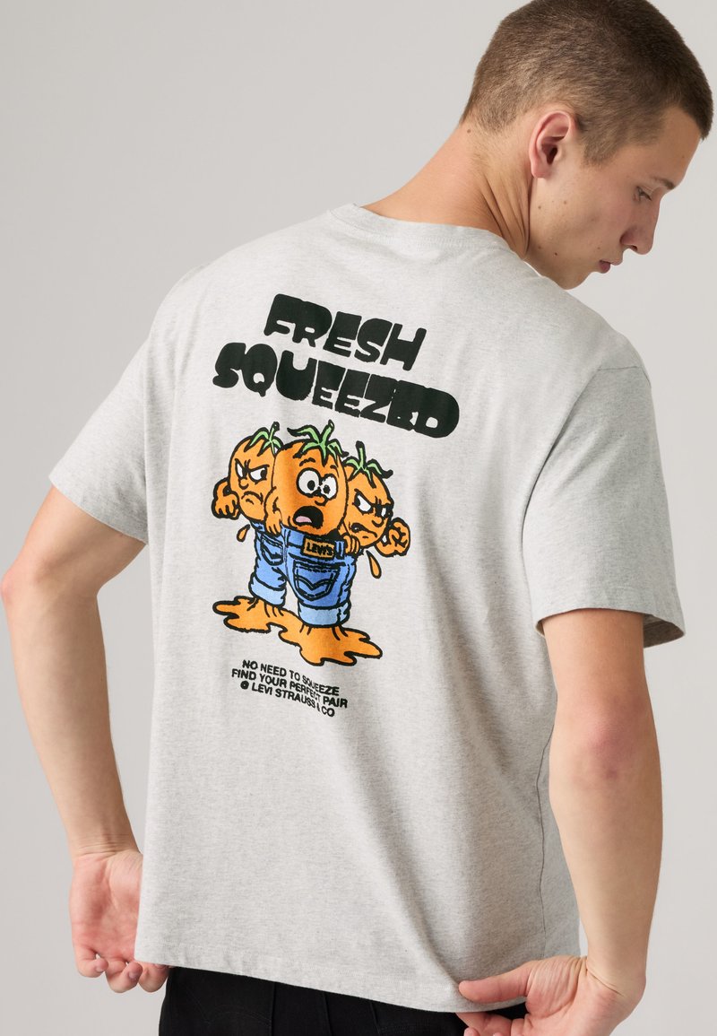 Mann trägt hellgraues T-Shirt mit Grafik von drei ausgepressten orangen Charakteren in Jeans und dem Text „FRESH SQUEEZED“ auf dem Rücken.