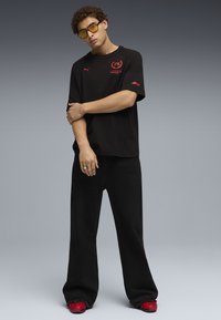 Mustat ylisuuret t-paidat, joissa on punainen logo, yhdistettynä mustiin leveälahkeisiin housuihin ja punaisiin kenkiin. Mallilla on päässään keltaiset aurinkolasit.