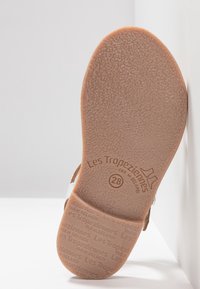 Hnědá gumová podrážka s texturovaným povrchem, vyražený název značky "Les Tropeziennes" a velikost "28," boční pohled odhaluje pásky z bílé a hnědé kůže.