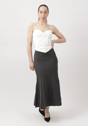 Kobieta stojąca, ubrana w biały, bezrękawnik w stylu gorsetu, długą ciemną, prążkowaną spódnicę, czarne szpilki, z jedną ręką na biodrze na jednolitym tle.